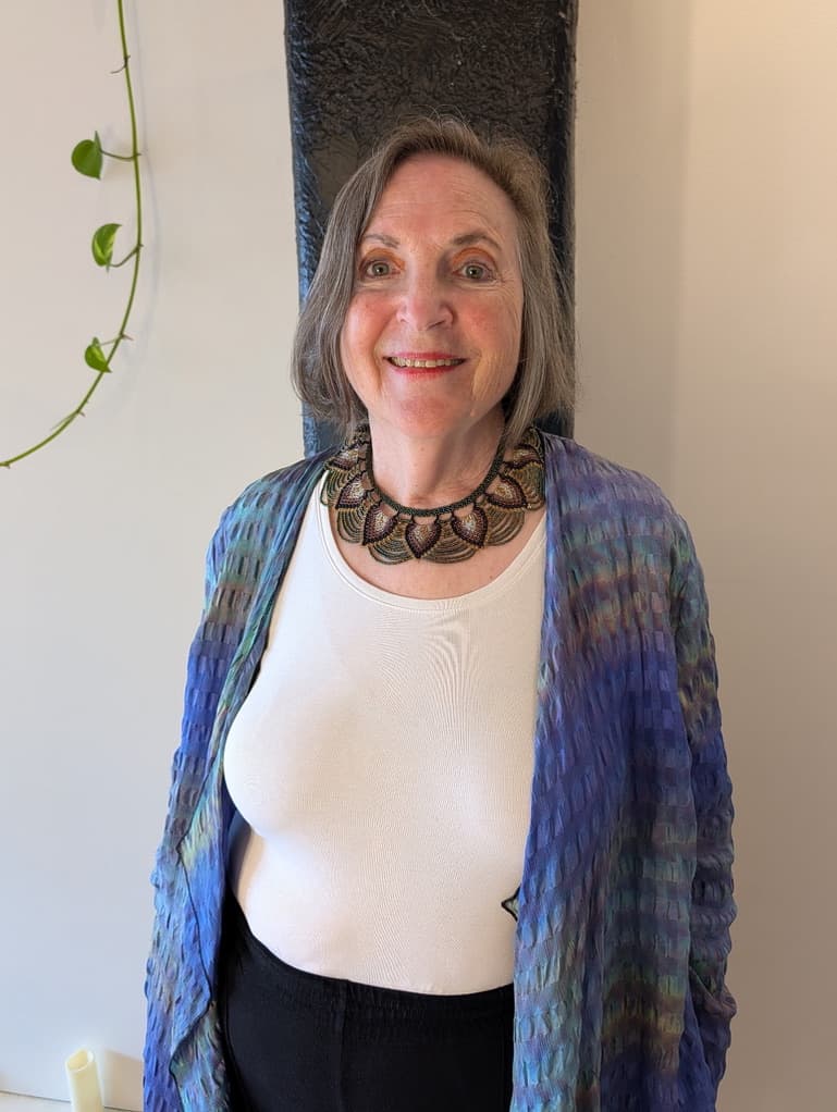 Smiling older woman with gray hair wearing a blue cardigan and an ornate beaded necklace.